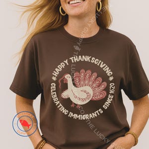 以下が含まれることがあります： 「Happy Thanksgiving Celebrating Immigrants Since 1620」と書かれた円形グラフィックが特徴の茶色のTシャツ。赤い肉垂と赤と白の羽の扇子を持つ七面鳥が描かれています。
