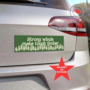 May include: Green bumper sticker with white text that reads "Strong winds make tough timber" and a row of white pine trees at the bottom. The sticker is on the back of a silver car. A red star sticker with white text that reads "Magnet or Sticker" is below the bumper sticker.