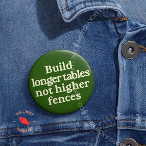 May include: A green circular badge with the white text "Build longer tables not higher fences". The badge is pinned to a blue denim jacket. The Lucky Guppy Co. logo is also visible.