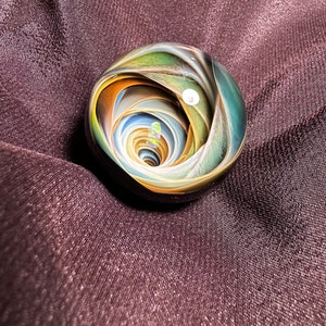 May include: A colorful glass marble with a swirling spiral design. The marble features shades of green, blue, yellow, and orange. The marble is set against a dark purple fabric background.