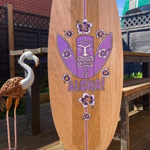 May include: A wooden surfboard with a purple tiki design, hibiscus flowers, and the word "Aloha!" in purple. The board has a light brown wood grain finish and a vertical purple stripe down the center.