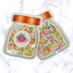 Pode incluir: Duas sacolas de plástico transparentes cheias de enfeites coloridos, variados, pequenos, decorativos, de resina e plástico. Os enfeites incluem fatias de frutas, estrelas, corações e pequenas formas redondas. As sacolas são etiquetadas com "Belle Rose".