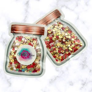 May include: Two clear, jar-shaped pouches filled with colorful sequins. The sequins are a mix of red, gold, and iridescent colors. One pouch has a label with the text "Belle Rose" and a floral design. The pouches have copper-colored lids.