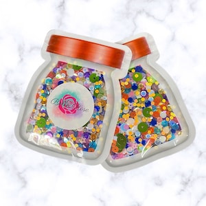 May include: Two clear plastic bags filled with colorful sequins and beads. The bags are shaped like jars with orange lids and a white label that says "Belle Rose".