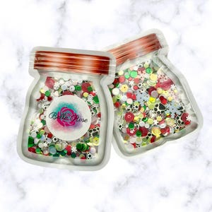May include: Two clear, jar-shaped pouches filled with colorful sequins. The sequins are in various shapes and colors, including red, green, white, and black. Each pouch has a copper-colored lid and a label with the text "Belle Rose".