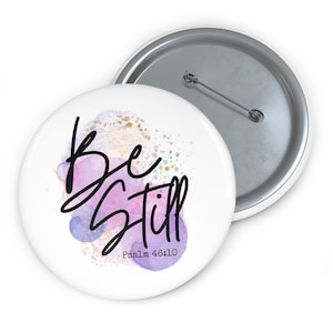 May include: A white button with a watercolor background in shades of purple and pink with gold glitter. The text "Be Still" is written in black cursive script with the words "Psalm 46:10" written below in black.