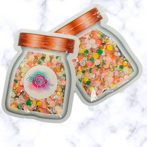 May include: Two clear plastic jars filled with colorful sprinkles and beads. The jars are labeled "Belle Rose".