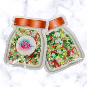 May include: Two clear plastic jars filled with colorful confetti and sequins. The jars are labeled "Belle Rose" and have a white background with a red, yellow, and blue flower design. The confetti and sequins are in various shapes and sizes, including stars, circles, and watermelon slices.