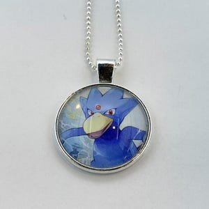 May include: A silver chain necklace with a round pendant featuring a blue and purple cartoon bird with red eyes. The bird is depicted with a white beak and is surrounded by a white background with blue and white swirls.