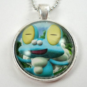 May include: A silver necklace with a round glass pendant featuring a cartoon image of a blue and yellow frog with a white background. The frog is smiling and has its eyes closed.