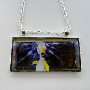 May include: Silver-toned rectangular pendant necklace featuring a cartoon character with yellow and white coloring. The pendant has a silver frame and a chain. The background of the pendant is a dark blue with white and purple rays.