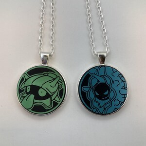 May include: Two silver-toned necklaces with round pendants. One pendant features a green cartoon character on a black background, the other a blue cartoon character on a black background. Both pendants have a silver border and are suspended from a delicate chain.