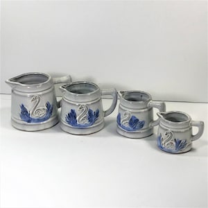 May include: Set of four white ceramic measuring cups with blue and white swan designs. The cups are different sizes and have a handle on the side.