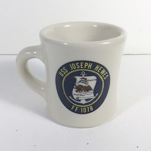 May include: White ceramic coffee mug with a blue and gold emblem that reads "USS JOSEPH HEWES FF 1078". The emblem features a ship with a bald eagle and an anchor.