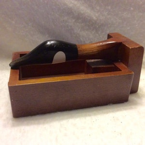 May include: Wooden duck-shaped nutcracker with a black head and a brown body. The nutcracker is in a wooden box with a hinged lid.