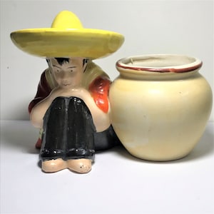 May include: A ceramic planter featuring a figurine of a person wearing a yellow sombrero and sitting with a large, cream-colored pot.