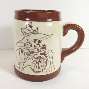 May include: A brown ceramic mug with a white glaze. The mug features a cartoon illustration of a man with a beard and a hat, holding a pipe in his mouth.