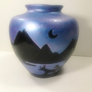 Op de afbeelding: Een blauwe keramische vaas met een zwart silhouet van bergen, een halve maan en een hert. De vaas heeft een glanzende afwerking.