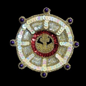 May include: A circular, gold-colored, beaded design with a red and gold anchor in the center. The design is surrounded by a ring of iridescent sequins and has eight purple beads around the outer edge.