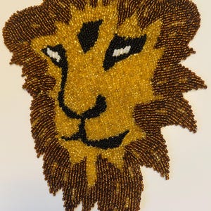 May include: A beaded lion face with a golden mane and face, brown accents, and black details. The lion's eyes are white. The artwork is likely handmade and suitable for wall decor or craft projects.