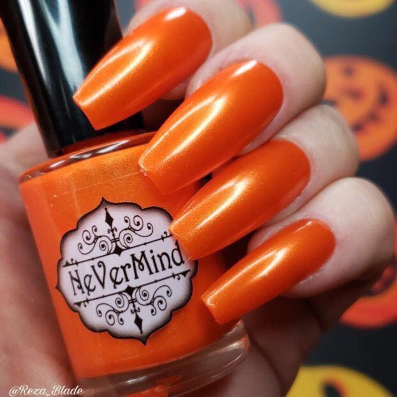 May include: Orange nail polish in a glass bottle with a black label that says "NeverMind". The bottle is being held by a hand with orange nail polish on the nails.