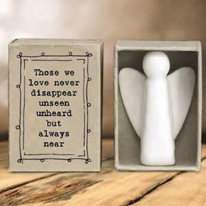 May include: A white ceramic angel figurine with outstretched wings, presented in an open matchbox. The adjacent box features the text "Those we love never disappear unseen unheard but always near" in black.