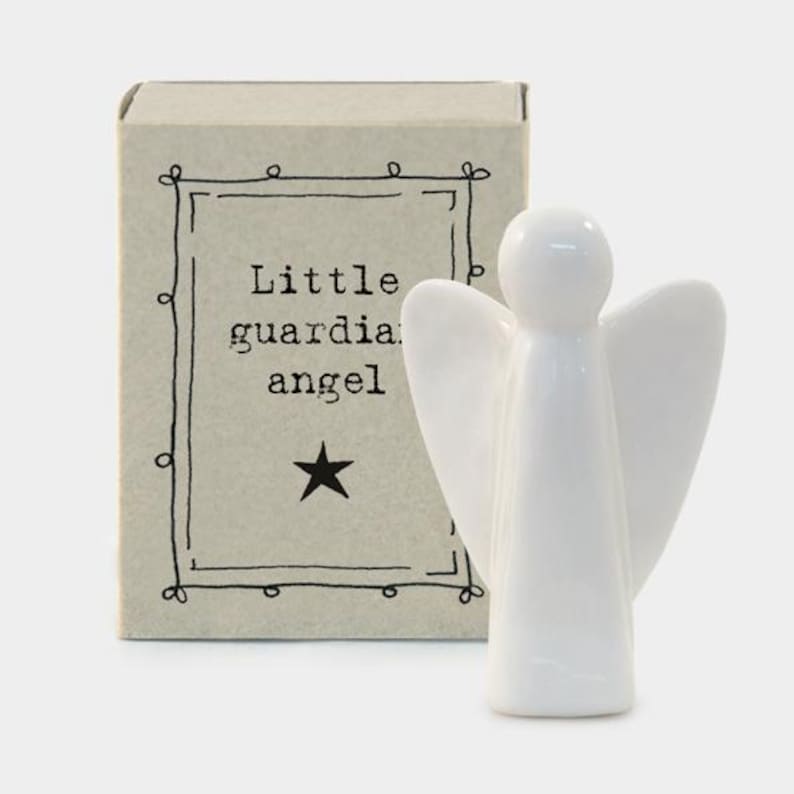 Puede incluir: Una peque&ntilde;a figura de &aacute;ngel de cer&aacute;mica blanca junto a una caja. La caja es beige con un marco negro y las palabras "Little guardian angel" y una estrella. El &aacute;ngel tiene alas extendidas.