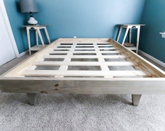 Puede incluir: Un marco de cama de madera con una base de listones y cuatro patas. El marco está hecho de madera sin terminar y tiene un color de madera natural.