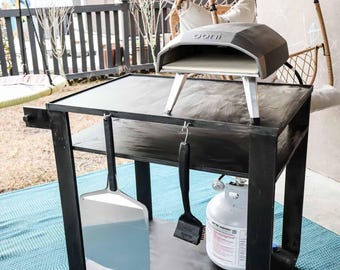 Könnte beinhalten: Ein schwarzer Metallwagen mit einer Holzplatte, einem Pizzaofen, einer Pizzaschaufel, einer Grillbürste und einer Propangasflasche. Der Wagen hat Räder und steht auf einem blau-weiß gemusterten Teppich.
