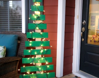 May include: A green wooden Christmas tree with white lights on a porch. The tree is made of stacked wooden planks and has a simple wooden base. The porch has a black and white chequered doormat with the text "Come back with tacos".