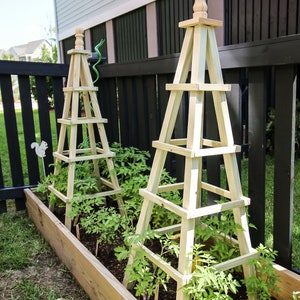 May include: Two wooden garden trellises with a light brown finish, supporting tomato plants in a raised garden bed. The trellises are made of square wooden slats and have a pointed top.