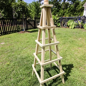 May include: A wooden garden trellis with a pointed top and four levels. The trellis is made of natural wood and is standing on a green lawn.