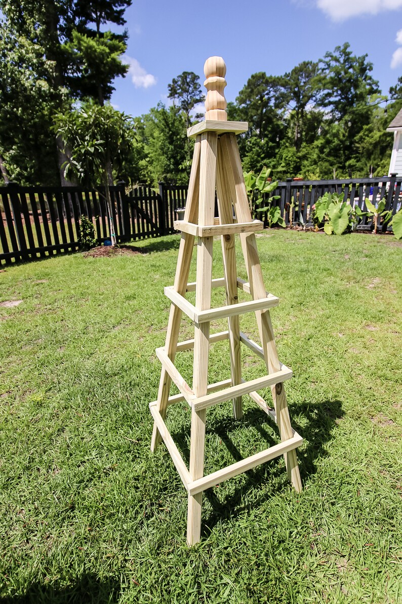 May include: A wooden garden trellis with a pointed top and four levels. The trellis is made of natural wood and is standing on a green lawn.
