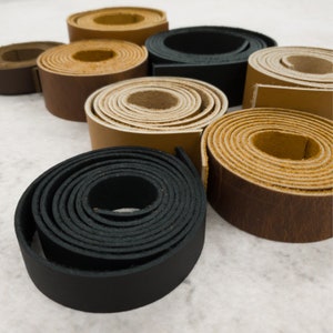 May include: Assortment of leather straps in various colors and widths, including black, brown, tan, and cream. The straps are rolled up and arranged in a semi-circle on a white surface.