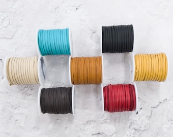 May include: A collection of spools of colorful suede cord in various colors, including turquoise, black, tan, brown, yellow, dark brown, and red. The cords are neatly wound on white spools, arranged against a textured white background.