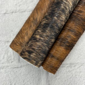 May include: Close-up of three rolled pieces of cowhide leather. The leather has a natural brindle pattern in shades of brown and black. The edges of the leather are visible, showing the thickness and texture of the material. The cowhide is likely used for crafting.