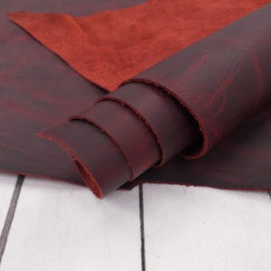 May include: Rolled burgundy leather with a rough texture. A piece of red leather is visible in the background. The leather has a distressed appearance, with variations in color and tone. The material appears to be high-quality and suitable for crafting.
