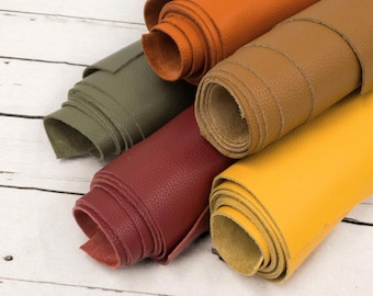 May include: A selection of rolled leather sheets in shades of orange, olive green, burgundy, tan, and yellow. The leather has a textured, pebbled finish. The rolls are displayed on a white wooden surface. Suitable for various craft projects.