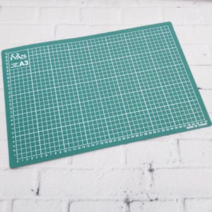 May include: A green A3 cutting mat with white grid lines and measurement markings. The mat has the text "VAN A3" in the upper left corner and "MADE IN TAIWAN" in the lower right corner. The mat is designed for crafting and measuring.