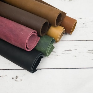 May include: A collection of rolled leather sheets in various colors including black, deep burgundy, forest green, mustard yellow, tan, and dark brown. Each roll displays a smooth, matte texture and consistent thickness, arranged in a diagonal stack.