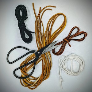 May include: A close-up of a set of leather cords in brown, white and black, with a pair of scissors. The cords are arranged in a starburst pattern, with the scissors in the centre.