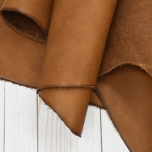 May include: Close-up of a folded piece of tan leather. The leather has a smooth, shiny surface with visible grain and a rough, unfinished edge. The background is a white wooden surface.