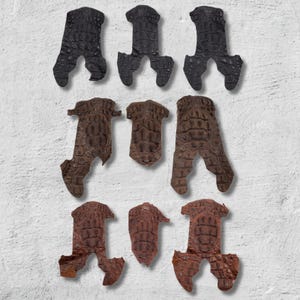 May include: Nine pieces of textured crocodile leather in various shades of brown and black. The pieces are cut into irregular shapes, some resembling feet or claws, and are arranged against a white background.