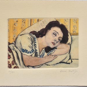 May include: A print of a woman asleep, depicted in gentle hues. She lies on a pillow, wearing a pale garment with blue floral details. The backdrop showcases a yellow floral design. Signed "Henri-Matisse".
