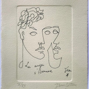 May include: A black line art print on cream-colored paper featuring two faces. The text "Les anges s'étonnent" is written below the faces, with the artist's signature and a number. The artwork is a minimalist style.