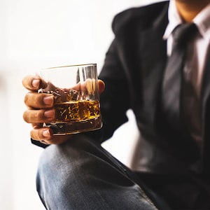 May include: A clear, patterned glass filled with amber liquid and ice cubes is held by a person wearing a black suit and tie. The glass has a gold rim. The person is wearing blue jeans.