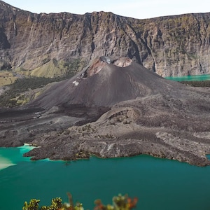 May include: A volcanic crater lake with a dark gray volcanic cone in the center. The lake is a vibrant turquoise blue and the surrounding landscape is a mix of dark gray volcanic rock and green vegetation.