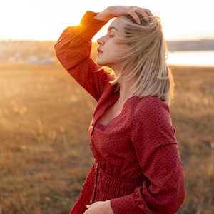 May include: A woman with blonde hair wearing a red dress with a white polka dot pattern stands in a field with her eyes closed and hand on her head. The sun is setting in the background.