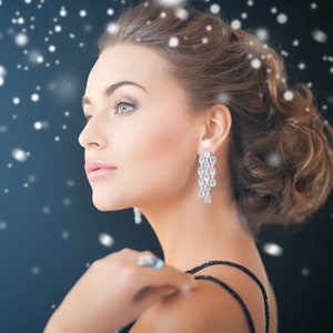 May include: A woman wearing elegant, dangling earrings with multiple clear, sparkling stones. The earrings are the focal point, reflecting light. The woman is wearing a black dress and has her hair styled up. The background is a dark blue with white dots.