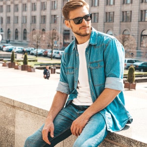 May include: A man wearing a light blue denim shirt over a white t-shirt and blue jeans. He is wearing sunglasses and sitting on a concrete ledge.